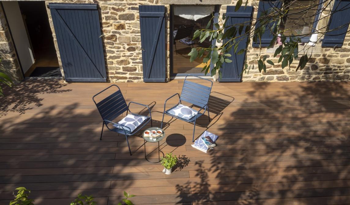 Terrasse en lames de bois composite teinte ipé avec salon de jardin bleu métallique, façade en pierre avec volets bleus, ombres d'arbres projetées sur le sol, aménagement extérieur élégant avec bois composite pour terrasse, idéal pour bardage et revêtement extérieur