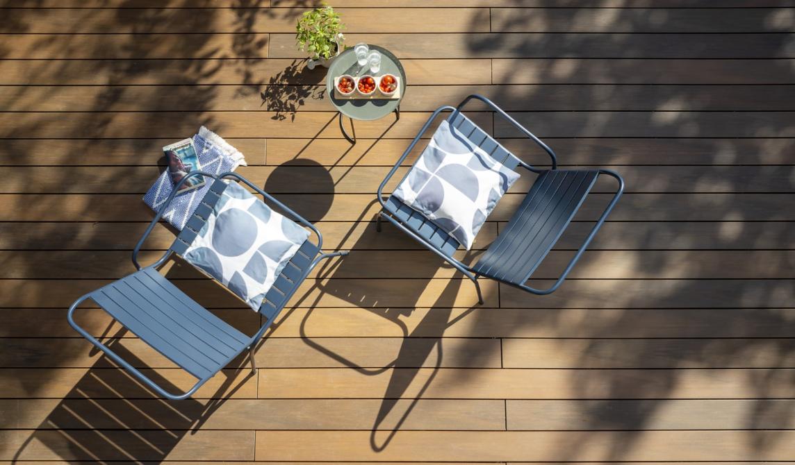 Vue plongeante sur terrasse en lames de bois composite teinte ipé avec deux chaises longues bleues à coussins motifs ronds, petite table ronde avec tomates et plante, ombres projetées sur les lames de bois composite pour terrasse, aménagement extérieur moderne et chaleureux