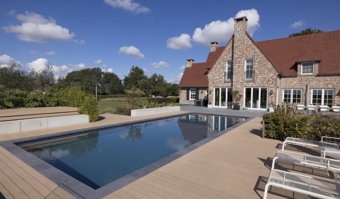 Élégante maison en pierre avec toit rouge, terrasse en bois composite entourant une piscine rectangulaire, mobilier extérieur moderne et jardin paysager en arrière-plan