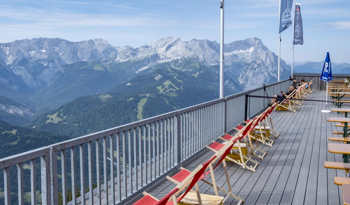 Terrasse panoramique en bois composite foncé avec garde-corps métallique, transats en bois avec toiles rouges alignés face à une vue spectaculaire sur les montagnes, drapeaux flottant au vent, point d'observation en altitude