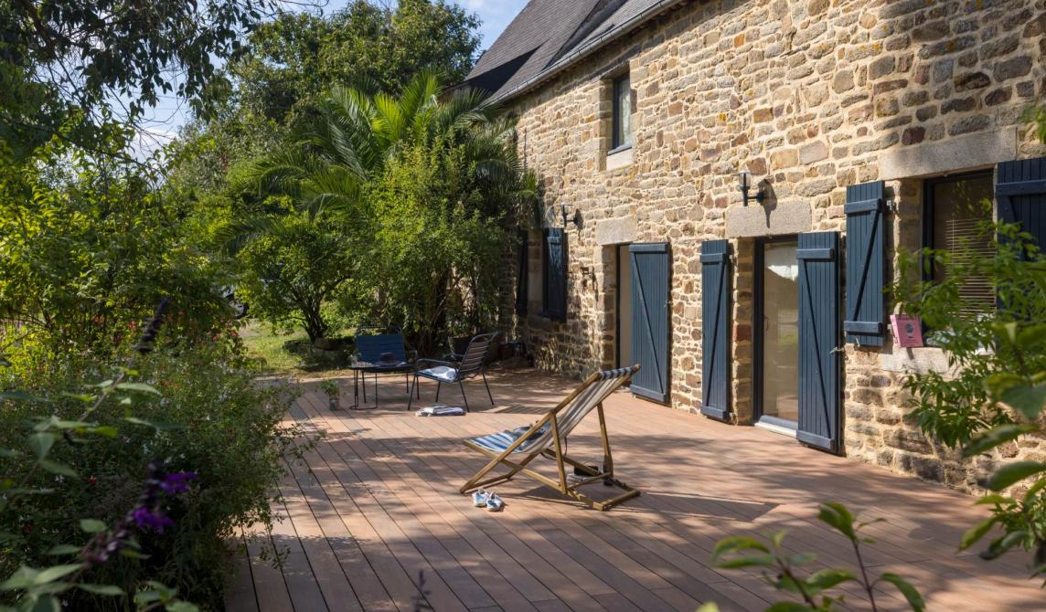 Grande terrasse en lames de bois composite teinte ipé entourant une maison bretonne en pierre avec volets bleus, transat et salon de jardin, végétation luxuriante, parfait exemple d'aménagement extérieur avec bois composite pour terrasse, alternative au bardage traditionnel