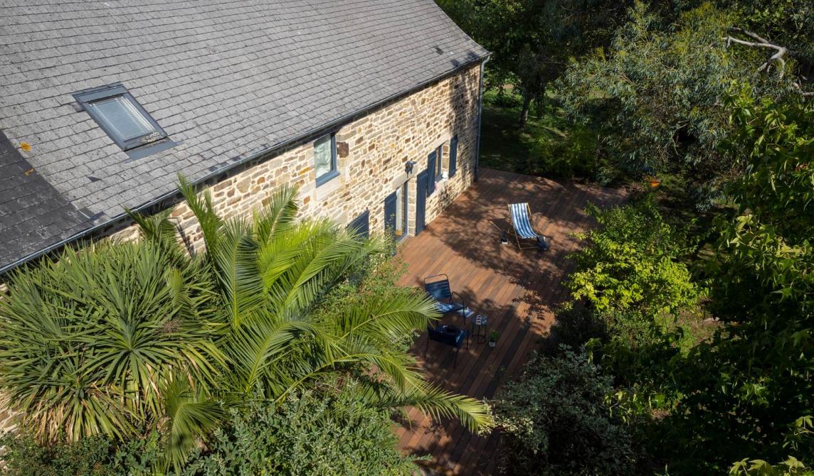 Vue aérienne d'une maison en pierre avec toiture en ardoise, terrasse en lames de bois composite teinte ipé entourée de végétation méditerranéenne et palmiers, salon de jardin bleu, parfaite intégration du bois composite pour terrasse dans un environnement naturel, alternative esthétique aux lames bois ipé traditionnelles