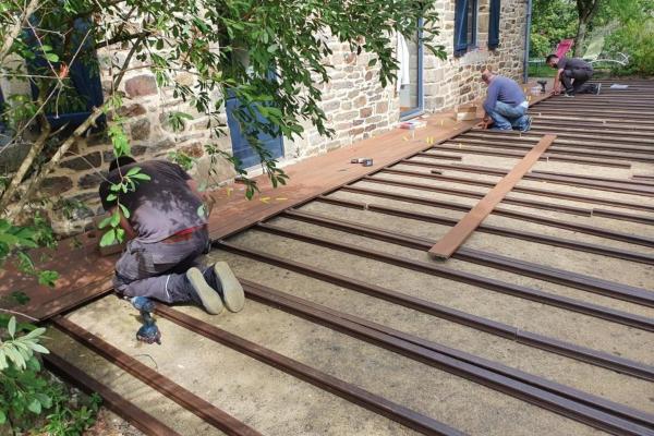 Installation d'une terrasse en bois composite Silvadec sur dalle béton, montrant la structure de lambourdes parallèles, avec un installateur au travail, des outils de pose et un mur en pierre ancien en arrière-plan