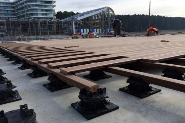 Installation de terrasse en bois composite Silvadec sur plots réglables en plastique noir, montrant la structure de lambourdes avant la pose complète des lames, sur un chantier de construction avec immeubles en arrière-plan