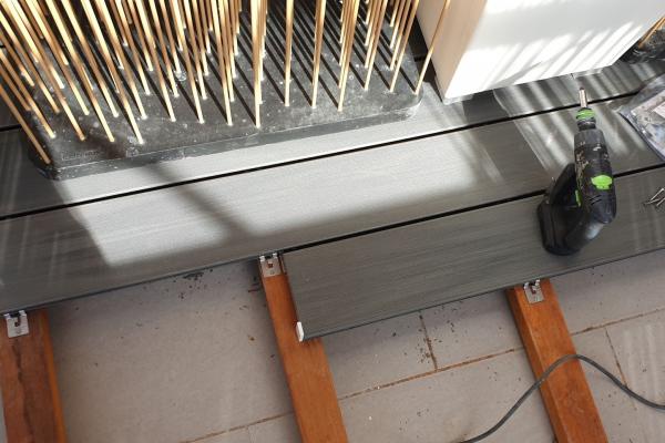 Système de fixation par clips pour lames de terrasse en bois composite, montrant les clips métalliques installés entre les lames et fixés sur les lambourdes en bois, avec une bouteille d'eau visible sur la terrasse en construction
