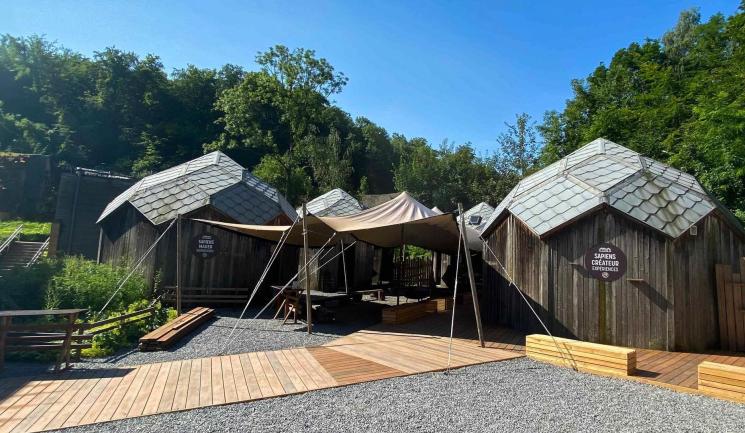 Wooden exhibition cabins with composite PRM ramp, slate roofs and shade sails, set in a landscaped area with gravel and vegetation